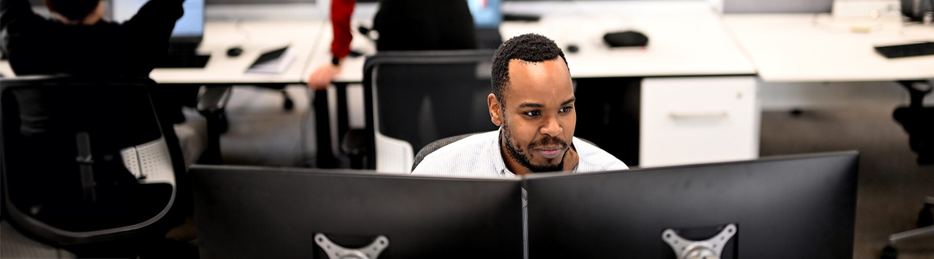 A man sits between computer monitors in a busy office, focusing intently on his screen.