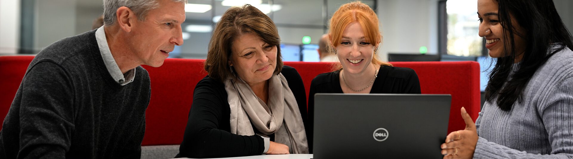 Four colleagues smiling and discussing over a laptop in a modern office setting.